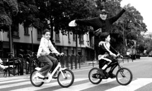 Cette photographie en noir et blanc met en scène trois personnes à vélo. Le sujet principal est l’homme au centre, levant les bras en équilibre sur sa roue arrière, créant un effet dynamique. La composition est équilibrée, avec les deux autres cyclistes en arrière-plan, ajoutant de la profondeur.Le cadrage est serré, centré sur les vélos et les personnages. La lumière est naturelle, probablement en journée, avec des ombres douces. Les couleurs dominantes, bien que monochromes, jouent sur les contrastes de gris et de noir.Le style est spontané et joyeux, évoquant une ambiance ludique et insouciante.
