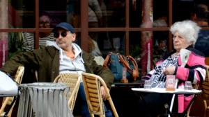 Cette photographie montre deux personnes âgées assises à une terrasse de café. Le sujet principal est ce couple, l’homme en veste verte et chapeau, la femme en veste rose, tous deux absorbés dans leurs pensées.La composition est simple et naturelle, avec un cadrage serré sur les deux individus. La lumière semble douce et naturelle, probablement en journée. Les couleurs dominantes sont sobres : tons verts, roses et gris, avec des touches de bois clair pour les tables et chaises.L’ambiance est calme et intimiste, évoquant une pause détendue dans un cadre urbain.
