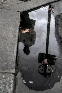 Cette photographie montre une personne vue de dessus à travers une flaque d’eau, reflétant son image inversée. Le sujet principal est le pied et la jambe de la personne, ainsi que son reflet déformé.La composition repose sur un cadrage serré et symétrique, centré sur la flaque. La lumière semble naturelle, avec des contrastes doux, créant des reflets sombres et des nuances claires.Les couleurs dominantes sont les tons gris et noirs, avec des touches de blanc et de bleu dans l’eau. Le style est réaliste et documentaire, évoquant une ambiance urbaine et mélancolique, presque poétique.