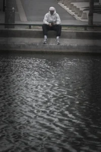 Cette photographie met en scène un skateboarder assis sur une margelle, les pieds dans l’eau. Le sujet principal est le jeune homme, vêtu d’un sweat et d’un bonnet, en position détendue.La composition est épurée, avec un cadrage serré sur le sujet et un premier plan dominé par l’eau. La lumière, douce et tamisée, crée des contrastes subtils. Les couleurs, en noir et blanc, renforcent l’ambiance mélancolique et introspective. Le style est documentaire et sobre, évoquant une atmosphère calme et contemplative.