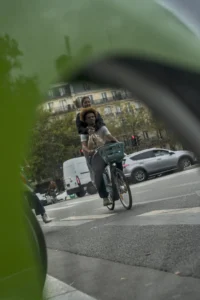 Cette photographie montre une personne à vélo comme sujet principal, vue à travers un rétroviseur ou un objectif déformant. La composition est dynamique, avec un cadrage serré et légèrement flou, accentuant le mouvement. La lumière semble naturelle, mais un peu tamisée, typique d’un jour nuageux.Les couleurs dominantes sont des tons froids et neutres (gris, bleu pâle), adoucis par des touches de vert (végétation) et de blanc (véhicules). Le style est documentaire et spontané, capturant une scène urbaine du quotidien. L’ambiance est réaliste et légèrement mélancolique, reflétant le rythme effréné de la ville.
