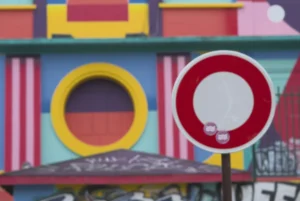 Cette photographie met en avant un **panneau de signalisation** rouge et blanc, partiellement flou, avec des cercles barrés, symbolisant une interdiction. En arrière-plan, des **structures colorées** aux teintes vives (rose, bleu, jaune, rouge) forment un décor ludique et enfantin, évoquant un parc d’attractions ou une aire de jeux.Le **cadrage** est serré, avec une **mise au point sélective** (effet bokeh), créant un contraste entre le premier plan net et l’arrière-plan flou. Les **couleurs dominantes** sont vives et saturées, apportant une ambiance **joyeuse et dynamique**. Le style est **urbain et artistique**, avec une touche de fantaisie. La lumière semble naturelle, bien répartie, sans ombre marquée.