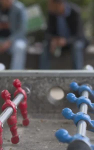 Cette photographie met en scène deux figurines stylisées représentant des molécules, l'une rouge et l'autre bleue, posées sur une surface en béton. Le sujet principal est ces figurines, évoquant une structure chimique.La composition est simple et épurée, avec un cadrage serré sur les figurines. L'arrière-plan flou montre des personnes, suggérant un environnement urbain ou public. La lumière naturelle semble douce, éclairant les objets sans ombres marquées. Les couleurs dominantes sont le rouge vif et le bleu électrique, contrastant avec le gris du béton.Le style est minimaliste et moderne, créant une ambiance à la fois scientifique et artistique.