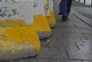 Cette photographie met en scène des **barrières de protection jaune et grise** posées sur un trottoir en pavés. Le **sujet principal** semble être ces barrières, partiellement endommagées.La **composition** est simple, avec un **cadrage serré** sur les obstacles, laissant peu de place à d'autres éléments. Un **pied en mouvement** (vêtu de baskets) apparaît en haut à droite, suggérant une personne marchant.Les **couleurs dominantes** sont le **jaune vif** des barrières et le **gris** des pavés et de la peinture écaillée. La **lumière** est naturelle, sans ombre marquée, créant une ambiance **neutre et urbaine**. Le style est **documentaire et réaliste**.