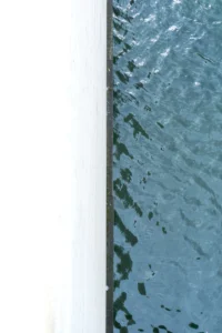 Cette photographie capture une portion de surface aquatique reflétant le ciel. Le **sujet principal** est l’eau, avec ses reflets bleutés et ses ondulations. La **composition** est minimaliste, avec un **cadrage serré** sur une bande verticale, séparant deux espaces visuels distincts.La **lumière** semble naturelle, douce et diffuse, créant des jeux de transparence. Les **couleurs dominantes** sont des teintes de bleu et de blanc, évoquant une ambiance calme et apaisante. Le **style** est épuré, presque abstrait, tandis que l’**ambiance** est contemplative et sereine, comme une invitation à observer la beauté simple des éléments naturels.