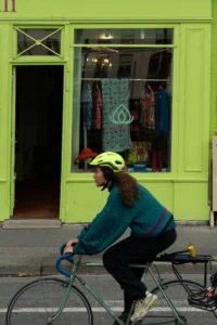 Cette photographie met en scène une personne à vélo comme sujet principal, vêtue d’une tenue sportive et d’un casque jaune. La composition montre un cadrage serré sur le cycliste, avec un arrière-plan de vitrine colorée reflétant des vêtements.Les couleurs dominantes sont le vert vif de la façade, le bleu du vêtement et le jaune du casque. La lumière semble naturelle, avec des ombres douces, créant une ambiance urbaine et dynamique. Le style est réaliste et spontané, capturant un moment de vie quotidienne dans un cadre urbain coloré et vivant.