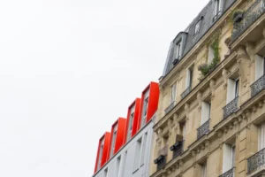 Cette photographie met en scène un immeuble parisien typique avec ses fenêtres et balcons en fer forgé. Le sujet principal est les trois structures verticales rouges et blanches, probablement des éléments architecturaux ou artistiques modernes, contrastant avec le bâtiment ancien.Le cadrage est serré, mettant l'accent sur ces éléments. La lumière est douce et diffuse, sans ombre marquée, créant une ambiance neutre et légèrement mélancolique. Les couleurs dominantes sont les tons beiges et gris de la façade, rehaussés par le rouge vif des structures. Le style est urbain et contemporain, mêlant tradition et modernité.