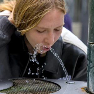 Cette photographie montre une jeune femme comme sujet principal, penchée pour boire à un jet d’eau. Le cadrage est serré, centré sur son visage et ses mains.La composition est dynamique, avec un premier plan occupé par l’eau et un arrière-plan flou. La lumière semble naturelle et douce, mettant en valeur les détails du visage et les éclaboussures. Les couleurs dominantes sont des tons froids (bleu grisâtre de l’eau, noir du vêtement) contrastant avec les teintes chaudes de la peau.Le style est documentaire et spontané, créant une ambiance authentique et vivante.