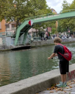 Cette photographie montre un enfant comme sujet principal, debout sur une berge, occupé à lancer une ligne de pêche dans un canal. La composition est équilibrée, avec l’enfant en premier plan et un pont vert en arrière-plan.Le cadrage est serré, mettant en valeur les détails de l’activité. La lumière est naturelle et douce, suggérant un moment en journée. Les couleurs dominantes sont les teintes vertes du pont, les tons bleus du canal et les couleurs vives du vêtement rouge de l’enfant. Le style est réaliste et spontané, créant une ambiance paisible et presque nostalgique.