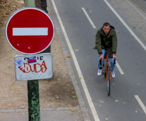 Cette photographie montre une personne à vélo comme sujet principal, roulant sur une piste cyclable. La composition met en avant un panneau "Stop" partiellement tagué avec le mot "Luda".Le cadrage est serré sur le vélo et le cycliste, avec un arrière-plan incluant une route et un trottoir. La lumière semble naturelle, probablement en journée, avec des ombres peu marquées. Les couleurs dominantes sont les tons gris (asphalte), vert (panneau), rouge (panneau Stop) et des touches de vert et brun (vêtements du cycliste).Le style est documentaire et réaliste, capturant une scène urbaine du quotidien. L’ambiance est calme et ordinaire.