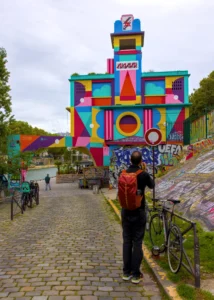 Cette photographie met en scène un **mur coloré et muraliste** aux formes géométriques vives, représentant une architecture futuriste. Le **sujet principal** est un homme en rouge, dos à la caméra, observant le site.La **composition** est dynamique, avec un **cadrage en contre-plongée** accentuant la hauteur du mur. Les **couleurs dominantes** sont vives : rose, bleu, jaune, vert et orange. La **lumière** est naturelle, légèrement voilée par un ciel nuageux, créant une ambiance lumineuse mais douce.Le **style** est urbain et artistique, évoquant une scène culturelle. L’**ambiance** est à la fois festive et contemplative, dans un cadre de rue animé.