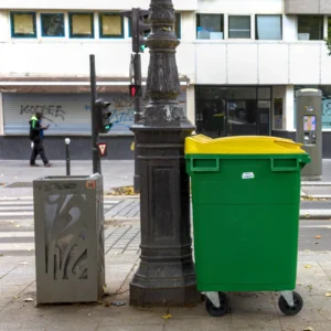 Cette photographie met en scène un **sujet urbain** avec pour éléments principaux un **poubelin graffití**, un **poteau ornemental** et un **bac de recyclage vert et jaune**. La composition est simple et symétrique, centrée sur ces objets.Le cadrage est serré, mettant en valeur les détails. La lumière semble naturelle, avec des ombres douces, suggérant un jour nuageux. Les **couleurs dominantes** sont les tons gris (bâtiments, poubelle), vert et jaune vif (bac), et noir (poteau).Le style est **documentaire et réaliste**, capturant une scène banale de la ville. L’ambiance est **urbaine et légèrement mélancolique**.