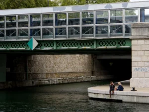 Cette photographie montre deux personnes assises sur une berge en béton, près d’un canal. Le sujet principal est ce duo en conversation, sous un pont métallique et vert.La composition est équilibrée, avec le pont en arrière-plan et les arbres en toile de fond. Le cadrage est serré sur les personnages et leur environnement immédiat. La lumière semble naturelle, avec des ombres douces.Les couleurs dominantes sont les verts du pont et de la végétation, les gris du béton et les bleus du canal. Le style est réaliste et documentaire, capturant une scène quotidienne. L’ambiance est calme et intimiste.