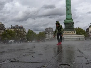Cette photographie met en scène une personne en mouvement, vêtue d’un manteau vert et de bottes rouges, marchant sous une pluie fine. Le sujet principal est centré, avec en arrière-plan une colonne monumentale (probablement la colonne Vendôme à Paris).La composition est équilibrée, avec un cadrage serré sur le sujet et une perspective légèrement basse. La lumière est tamisée par les nuages, créant une ambiance grise et mélancolique. Les couleurs dominantes sont les tons froids (gris, bleu) adoucis par les touches de vert et rouge du manteau.Le style est réaliste et documentaire, évoquant une atmosphère calme et contemplative.