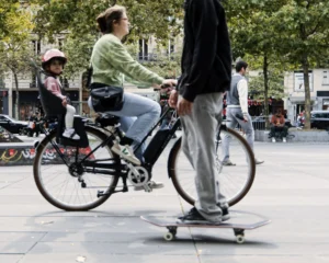 Cette photographie met en scène deux personnes en mouvement dans un espace urbain. Le sujet principal est un adulte sur un vélo avec un enfant assis devant lui, tandis qu’un autre adulte fait du skateboard à l’avant-plan.La composition est dynamique, avec un cadrage serré sur les jambes et les pieds des protagonistes, créant un effet de profondeur. La lumière semble naturelle, probablement en journée, avec des ombres douces. Les couleurs dominantes sont neutres et naturelles : gris (pavés, vêtements), vert (végétation), et touches de couleurs vives sur les vêtements et le vélo.L’ambiance est moderne et décontractée, reflétant une vie urbaine active.