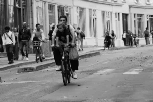Cette photographie en noir et blanc met en scène un cycliste comme sujet principal, transportant un sac volumineux. La composition montre une rue urbaine animée, avec des passants en arrière-plan.Le cadrage est serré sur le cycliste, mais inclut une partie de l’environnement. La lumière est naturelle, douce, créant des contrastes modérés. L’absence de couleurs accentue les textures et les jeux d’ombres.Le style est documentaire, réaliste, capturant un instant de vie quotidienne. L’ambiance est dynamique, reflétant le mouvement et l’agitation d’une ville.