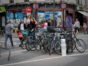 Cette photographie capture une scène urbaine animée. Le **sujet principal** est un groupe de vélos stationnés en bordure de trottoir, avec des personnes en arrière-plan.La **composition** est dynamique, avec un cadrage serré sur les vélos et une perspective légèrement basse. Les **couleurs dominantes** sont neutres (gris, noir des vélos) rehaussées par des touches vives (vêtements colorés des passants).La **lumière** semble naturelle, probablement diurne, avec des ombres douces. L’ambiance est **vivante et quotidienne**, reflétant une ville active et multiculturelle. Le style est **documentaire**, réaliste et sans artifice.