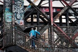Cette photographie montre un homme en bleu, sujet principal, marchant sous une pluie fine sur un pont métallique couvert de graffitis. La composition met en valeur les structures industrielles rouillées et les escaliers en métal. Le cadrage est serré, centré sur l’homme et les détails architecturaux.La lumière est tamisée, créant une ambiance grise et mélancolique. Les couleurs dominantes sont les tons froids (bleu, gris, rouille) et les touches de graffiti coloré. Le style est réaliste et documentaire, évoquant une atmosphère urbaine et poétique, presque onirique.