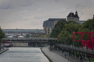 Cette photographie montre un pont métallique enjambant un canal urbain. Le sujet principal est ce pont et l’architecture environnante, incluant un bâtiment moderne et une tour historique à l’arrière-plan.La composition est équilibrée, avec le pont au centre et des arbres à droite. Le cadrage est large, capturant une vue panoramique. La lumière est douce, légèrement voilée par un ciel couvert, créant une ambiance calme.Les couleurs dominantes sont les tons gris et verts, adoucis par des touches de rouge et de bleu. Le style est réaliste et documentaire, évoquant une ambiance paisible et légèrement mélancolique.