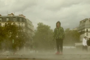 Cette photographie met en scène une personne vêtue d’un manteau vert et d’un chapeau, debout au centre d’une rue embrumée. La composition est symétrique, avec le sujet légèrement isolé par la brume.Le cadrage est serré sur le personnage, avec des bâtiments et des arbres en arrière-plan. La lumière est diffuse, atténuée par le brouillard, créant une atmosphère mystérieuse et mélancolique. Les couleurs dominantes sont les tons grisâtres et verts, renforçant une ambiance froide et onirique. Le style rappelle un réalisme poétique, évoquant une scène presque surnaturelle.