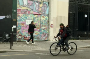 Cette photographie montre un cycliste comme sujet principal, vêtu d’un manteau sombre et d’un sac à dos, en mouvement. La composition inclut aussi deux personnes en arrière-plan près d’un mur couvert de graffitis colorés et variés.Le cadrage est serré sur le cycliste, avec une profondeur de champ qui met en valeur les détails du premier plan et les éléments urbains en arrière-plan. La lumière semble naturelle, un peu grise, typique d’un jour couvert. Les couleurs dominantes sont les teintes vives des graffitis contrastant avec les tons neutres de la ville.L’ambiance est urbaine et dynamique, mêlant art de rue et vie quotidienne.