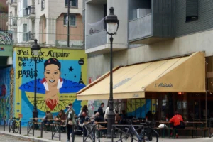Cette photographie montre une rue parisienne animée. Le **sujet principal** est un **mural coloré** représentant une femme souriante avec des éléments stylisés évoquant Paris. La **composition** associe ce mural à un café-restaurant nommé "L'Effervescence", avec ses tables et parasols jaunes.Le **cadrage** est serré sur le mural et l’établissement, incluant quelques passants et vélos. La **lumière** est naturelle, douce, typique d’une journée ensoleillée. Les **couleurs dominantes** sont vives : bleu, jaune, vert et rouge.Le **style** est urbain et artistique, mêlant street art et ambiance parisienne. L’**ambiance** est chaleureuse et dynamique, reflétant le charme typique des quartiers animés de Paris.