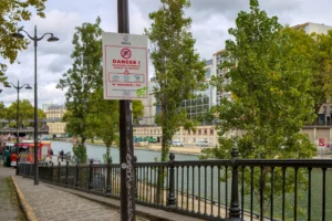 Cette photographie montre un panneau "Danger" indiquant une interdiction de stationner, fixé à un réverbère. La composition inclut une promenade bordée de rails noirs, des arbres en fleurs et un canal en arrière-plan. Le cadrage est large, capturant un espace urbain paisible.La lumière est naturelle, légèrement voilée par des nuages, créant une ambiance douce. Les couleurs dominantes sont les verts des arbres, les gris des bâtiments et du ciel, et les touches de rouge et blanc du panneau. Le style est réaliste et documentaire, évoquant une ambiance calme et urbaine.