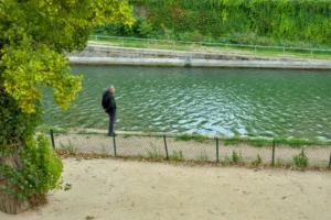 Cette photographie montre une personne en silhouette près d’un plan d’eau, observant le paysage. La composition est aérée, avec un cadrage en plan large incluant une berge herbeuse et des arbres.La lumière est naturelle et douce, suggérant un jour ensoleillé. Les couleurs dominantes sont les verts (eau, végétation) et les tons beiges (sable). Le style est épuré et réaliste, avec une ambiance calme et contemplative, évoquant la sérénité et la connexion avec la nature.