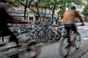 Cette photographie capture un cycliste en mouvement devant un parking à vélos. Le **sujet principal** est le cycliste, flou par effet de vitesse, créant un **style dynamique**.La **composition** montre une rue urbaine avec des vélos alignés et des bâtiments en arrière-plan. Le **cadrage** est serré, mettant l’accent sur le mouvement et l’avant-plan.Les **couleurs dominantes** sont neutres (gris, bleu clair) avec des touches de vert des arbres. La **lumière** semble naturelle, probablement en journée, avec des ombres douces. L’**ambiance** est urbaine et animée, reflétant la vie quotidienne en ville.