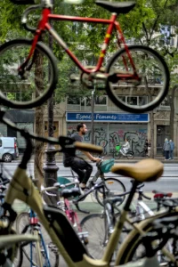Cette photographie met en scène un cycliste en mouvement, flou, au premier plan. La composition montre une scène urbaine avec des vélos stationnés en avant et un fond de rue animée. Le cadrage est serré, avec une perspective légèrement basse, accentuant la dynamique du sujet.Les couleurs dominantes sont les tons froids (gris, bleu) et chauds (rouge du vélo, vert des arbres). La lumière semble naturelle, avec des contrastes modérés. Le style est documentaire et spontané, capturant un instant de vie quotidienne. L’ambiance est vivante et urbaine, mêlant mouvement et immobilité.