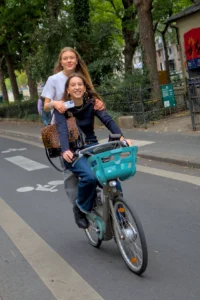 Cette photographie montre deux jeunes femmes sur un vélo en ville. Le sujet principal est la personne conduisant, avec sa passagère assise derrière elle dans un panier coloré.La composition est dynamique, avec un cadrage serré sur les deux femmes. La lumière est naturelle et semble provenir d’un jour ensoleillé, créant des ombres douces. Les couleurs dominantes sont les tons verts des arbres, le bleu du panier et les vêtements sombres des femmes. Le style est spontané et joyeux, évoquant une ambiance décontractée et urbaine.