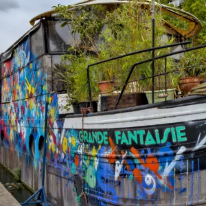 Cette photographie montre un bateau nommé **« Grande Fantaisie »**, décoré de fresques colorées et fantaisistes. Le sujet principal est ce bateau artistiquement peint, avec des motifs variés et joyeux.La composition met en valeur les couleurs vives et les détails du graffiti, encadrés par une végétation luxuriante. Le cadrage est serré, capturant une partie du bateau et son environnement immédiat. La lumière naturelle éclaire la scène, mettant en relief les teintes vives et les contrastes.Les couleurs dominantes sont le bleu, le jaune, le vert et le rouge, créant une ambiance festive et créative. Le style est artistique et décalé, évoquant une touche bohème et libre.