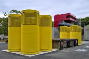 Cette photographie montre trois grands cylindres jaunes empilés, probablement des poubelles industrielles, placées sur un parking. En arrière-plan, on aperçoit des conteneurs métalliques et une structure rouge.La composition est épurée, avec un cadrage serré sur les cylindres. La lumière est naturelle, diffuse, sans ombres marquées, suggérant un jour nuageux. Les couleurs dominantes sont le jaune vif des cylindres et le gris des surfaces environnantes, avec une touche de rouge en arrière-plan.Le style est documentaire, réaliste, et l’ambiance industrielle et utilitaire.
