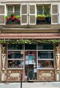 Cette photographie montre une boutique aux allures vintage. Le sujet principal est la façade d’un commerce avec ses grandes fenêtres et ses plantes en pots. La composition est symétrique, avec des éléments décoratifs comme des motifs en bois et des enseignes colorées.Le cadrage est serré, mettant en valeur les détails architecturaux. La lumière est naturelle et douce, éclairant les tons chauds. Les couleurs dominantes sont les bruns, verts et touches de rouge.Le style évoque un charme rétro et artisanal, créant une ambiance chaleureuse et accueillante.