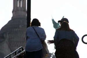 Cette photographie montre deux personnes dos à l'objectif, observant un monument flou en arrière-plan, probablement la Statue de la Liberté. La composition met en valeur leurs silhouettes et leurs gestes, l’une tenant un appareil photo. Le cadrage est serré, centré sur les sujets et leur environnement immédiat. La lumière est douce et diffuse, créant une ambiance légèrement brumeuse. Les couleurs dominantes sont des tons neutres (gris, bleu clair) avec des contrastes atténués. Le style est documentaire et naturel, évoquant une scène du quotidien, empreinte de calme et de contemplation.