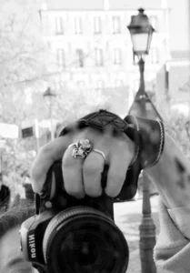 Cette photographie en noir et blanc met en scène une personne de dos, tenant un appareil photo. Le sujet principal est ses mains, dont l’une porte une bague ornée, suggérant une attention aux détails.La composition est centrée sur les mains et l’appareil, avec un cadrage serré. En arrière-plan, une rue avec des bâtiments et une lampe à gaz ajoute une touche urbaine et vintage. La lumière douce et contrastée accentue les textures et les ombres, créant une ambiance intime et nostalgique. Le style est documentaire et artistique, renforcé par le flou des éléments secondaires.