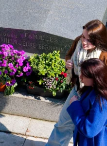 Cette photographie montre deux femmes devant un monument commémoratif. Le sujet principal est le mémorial dédié aux familles des jeunes du Béarn, avec des inscriptions gravées et des fleurs fraîches (pétunias et géraniums) en premier plan.La composition est symétrique, avec les femmes encadrant le monument. Le cadrage est serré sur les fleurs et les visages partiellement visibles. La lumière est naturelle et douce, probablement en journée. Les couleurs dominantes sont les verts des plantes, les roses et magentas des fleurs, et les tons neutres du monument.L’ambiance est à la fois solennelle et apaisante, évoquant le souvenir et la sérénité.