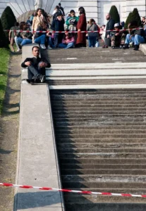 Cette photographie montre une personne assise sur une corniche étroite, dominant un escalier en pierre. Le sujet principal est cette personne, en position vulnérable et contemplative.La composition oppose la fragilité du sujet aux masses humaines en arrière-plan, séparées par une barrière. Le cadrage est serré sur la personne et l'escalier, avec une perspective dramatique.La lumière semble naturelle, légèrement froide. Les couleurs dominantes sont les tons gris (mur, pierre) et les teintes sombres des vêtements, avec des touches de rouge et bleu dans les détails (barrière, vêtements des spectateurs).L’ambiance est à la fois angoissante et contemplative, mêlant danger et solitude.