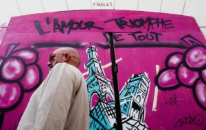 Cette photographie montre un homme âgé passant devant un mur tagué. Le sujet principal est le graffiti coloré, avec des inscriptions en français ("L’amour triomphe de tout") et des illustrations stylisées, dont la Tour Eiffel. La composition juxtapose l’art urbain et la réalité quotidienne. Le cadrage est serré sur le mur, avec l’homme en arrière-plan. Les couleurs dominantes sont le rose vif et le bleu, créant un contraste dynamique. La lumière semble naturelle, mettant en valeur les détails du graffiti. L’ambiance est à la fois vibrante et mélancolique, mêlant énergie artistique et passage du temps.
