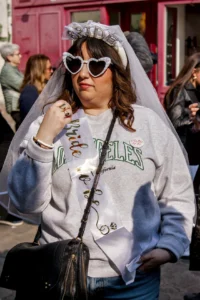 Cette photographie met en scène une femme comme sujet principal, vêtue d’un sweat à capuche et coiffée d’un voile de mariée stylisé, avec des lunettes à paillettes. Le cadrage est serré, centré sur elle, avec un arrière-plan flou montrant une rue animée.La lumière est naturelle et douce, mettant en valeur les détails de son visage et de ses accessoires. Les couleurs dominantes sont les tons pastel (blanc, gris) et des touches de rose et bleu. Le style est décalé et festif, évoquant une ambiance ludique et créative, presque carnavalesque.