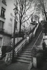 Cette photographie en noir et blanc met en scène un escalier en pierre bordé de rampes métalliques, sujet principal. La composition montre deux personnes en haut des marches, presque discrètes.Le cadrage est serré, encadrant l’escalier et ses abords urbains. La lumière, douce et légèrement tamisée, crée des contrastes subtils entre ombres et clartés, accentuant l’ambiance nostalgique.Les tons dominants, gris et noirs, renforcent une atmosphère mélancolique et intemporelle. Le style, sobre et documentaire, évoque une scène de vie quotidienne dans un quartier ancien, avec une touche poétique.
