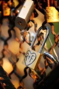 Cette photographie met en scène un ** cadenas en forme de cœur** avec les initiales "PF", accroché parmi d'autres cadenas. Le **sujet principal** est ce cadenas, symbolisant l’amour.La **composition** est serrée, avec un **cadrage rapproché** qui isole partiellement le cadenas. La **lumière** est douce et naturelle, créant des reflets dorés et des ombres douces. Les **couleurs dominantes** sont des tons chauds (or, bronze) et des touches de vert et de gris.Le **style** est réaliste et photographique, évoquant une **ambiance romantique et nostalgique**, typique des "ponts des amoureux".