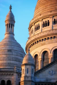 Cette photographie met en scène la **Sacré-Cœur de Montmartre**, avec ses **dômes blancs** comme sujet principal. La composition joue sur les **courbes architecturales** et les jeux d’ombres. Le cadrage serré isole les dômes, créant une perspective dynamique.La lumière, douce et dorée, suggère un **coup de soleil** en fin de journée, adoucissant les contours. Les **couleurs dominantes** sont des tons **beiges et blancs**, rehaussés par le bleu profond du ciel.Le style est **classique et monumental**, évoquant une ambiance **sereine et spirituelle**, typique des lieux emblématiques de Paris.