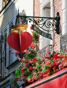 Cette photographie met en scène un balcon parisien orné de manière romantique. Le sujet principal est un cœur rouge suspendu à une balustrade en fer forgé, décorée de fleurs rouges et vertes. La composition est symétrique et harmonieuse, avec un cadrage serré sur le balcon.La lumière naturelle éclaire la scène, mettant en valeur les détails. Les couleurs dominantes sont le rouge vif des fleurs et du cœur, contrastant avec le noir de la balustrade et le vert des feuilles. Le style évoque un charme romantique et traditionnel, typique des décors parisiens. L’ambiance est chaleureuse et poétique.