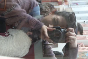Cette photographie montre une personne prenant une photo avec un appareil reflex. Le sujet principal est flou, mis en valeur par un effet de double exposition ou de miroir.La composition est simple, avec un cadrage serré sur les mains et l'appareil. La lumière semble naturelle, bien que légèrement tamisée. Les couleurs dominantes sont des tons chauds et neutres, avec des touches de rouge et de gris.Le style est documentaire et spontané, évoquant une ambiance intime et un peu mystérieuse. L'image suggère une activité créative ou un moment de capture improvisé.