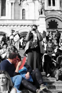 Cette photographie montre une scène urbaine animée. Le sujet principal semble être un homme assis par terre, en train de manger un sandwich, tandis qu'autour de lui, des personnes sont assises ou debout sur des marches.La composition est dynamique, avec un cadrage serré sur les individus. La lumière est naturelle, mais contrastée, créant des ombres marquées. Les couleurs dominantes sont des tons gris et sépia, donnant un aspect vintage à l'image.L'ambiance est celle d'une foule en mouvement, dans un lieu public, probablement un monument ou une place emblématique. Le style est documentaire et spontané.