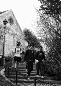 Cette photographie en noir et blanc montre trois jeunes hommes comme sujets principaux, descendant un escalier métallique devant une vieille bâtisse.La composition met en valeur les marches et la structure architecturale en arrière-plan. Le cadrage est serré sur les personnages, avec une profondeur de champ qui accentue les détails de l’escalier et des arbres environnants.La lumière semble naturelle, douce et légèrement tamisée, créant des contrastes subtils. Les couleurs dominantes, bien que monochromes, jouent sur les nuances de gris et de noir.Le style est documentaire et spontané, évoquant une ambiance calme et introspective.