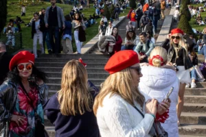 Cette photographie capture un groupe de personnes en extérieur, probablement lors d’un événement festif. Le sujet principal semble être les trois femmes en premier plan, vêtues de tenues colorées et coiffées de chapeaux rouges, évoquant une ambiance folklorique ou carnavalesque.La composition montre un escalier en pierre avec une foule en arrière-plan, créant une profondeur. Le cadrage est serré sur les femmes, mais inclut aussi des éléments contextuels. La lumière est naturelle et claire, suggérant une journée ensoleillée. Les couleurs dominantes sont vives : rouge, blanc, et des teintes pastel, renforçant l’ambiance joyeuse et festive. Le style est documentaire et spontané, reflétant une scène authentique et animée.
