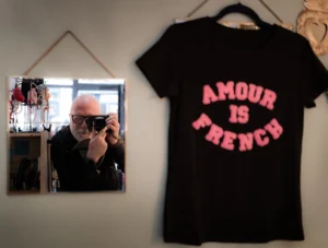 Cette photographie montre un t-shirt noir accroché avec l'inscription "Amour is French" en lettres roses. À gauche, un miroir reflète une personne prenant une photo avec un appareil argenté.La composition est simple et symétrique, avec un cadrage serré sur les éléments principaux. La lumière semble naturelle et douce, éclairant uniformément la scène. Les couleurs dominantes sont le noir du t-shirt, le rose des lettres, et les tons neutres du fond.Le style est minimaliste et décontracté, évoquant une ambiance intime et quotidienne.