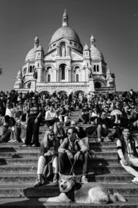 Cette photographie en noir et blanc capture la **Sacré-Cœur de Montmartre** à Paris. Le **sujet principal** est la basilique, avec ses dômes et sa façade imposante. La **composition** montre une foule sur les marches, créant un contraste entre l’architecture et les visiteurs.Le **cadrage** est serré sur les marches, mettant en valeur les gens et les animaux. La **lumière** est vive, avec des ombres marquées, typique d’un jour ensoleillé. Les **couleurs dominantes** sont les tons de gris, accentuant les contrastes.Le **style** est documentaire et réaliste, évoquant une **ambiance** vivante et populaire, mêlant spiritualité et quotidien.